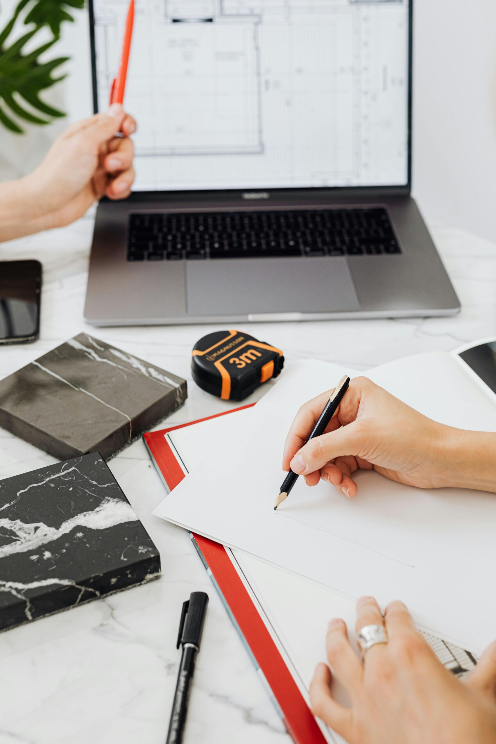 Close-up of hands sketching designs with laptop and tile samples in an office.