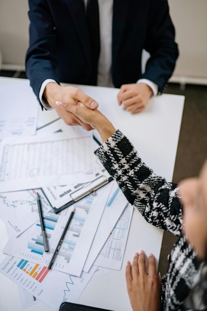 A close-up photo of a business handshake over financial documents in an office environment.